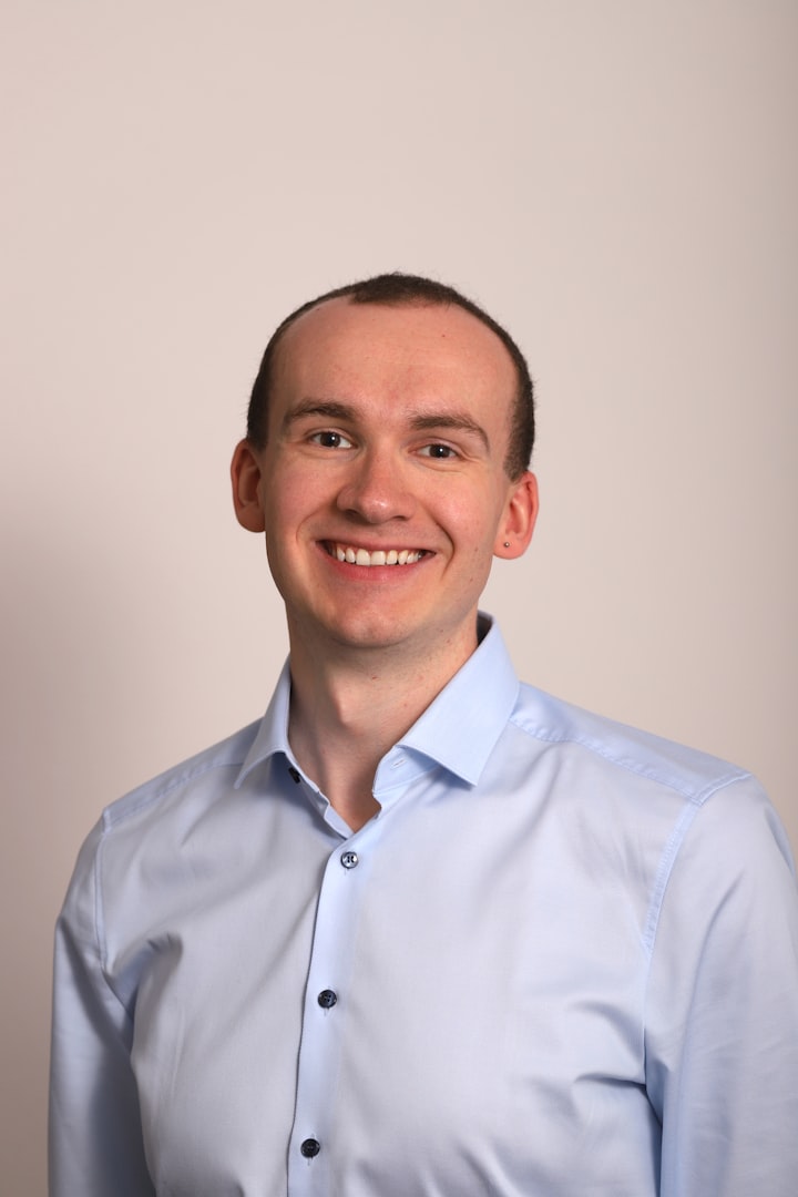 A man in a light blue polo shirt standing in front of a shelf filled with various kitchen items and textiles. The background is softly blurred, drawing focus to his friendly expression and casual style.